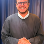 O Diácono Andrey Reimer de Melo sorri suavemente, com as mãos entrelaçadas, diante da nave principal do Santuário de Nossa Senhora do Rocio em Paranaguá, vestindo a túnica de sua ordenação. A luz suave do entardecer entra pelas janelas, destacando a arquitetura sacra e a aura de espiritualidade."