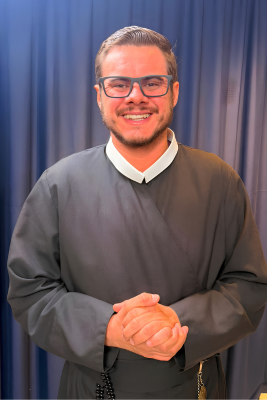 O Diácono Andrey Reimer de Melo sorri suavemente, com as mãos entrelaçadas, diante da nave principal do Santuário de Nossa Senhora do Rocio em Paranaguá, vestindo a túnica de sua ordenação. A luz suave do entardecer entra pelas janelas, destacando a arquitetura sacra e a aura de espiritualidade."