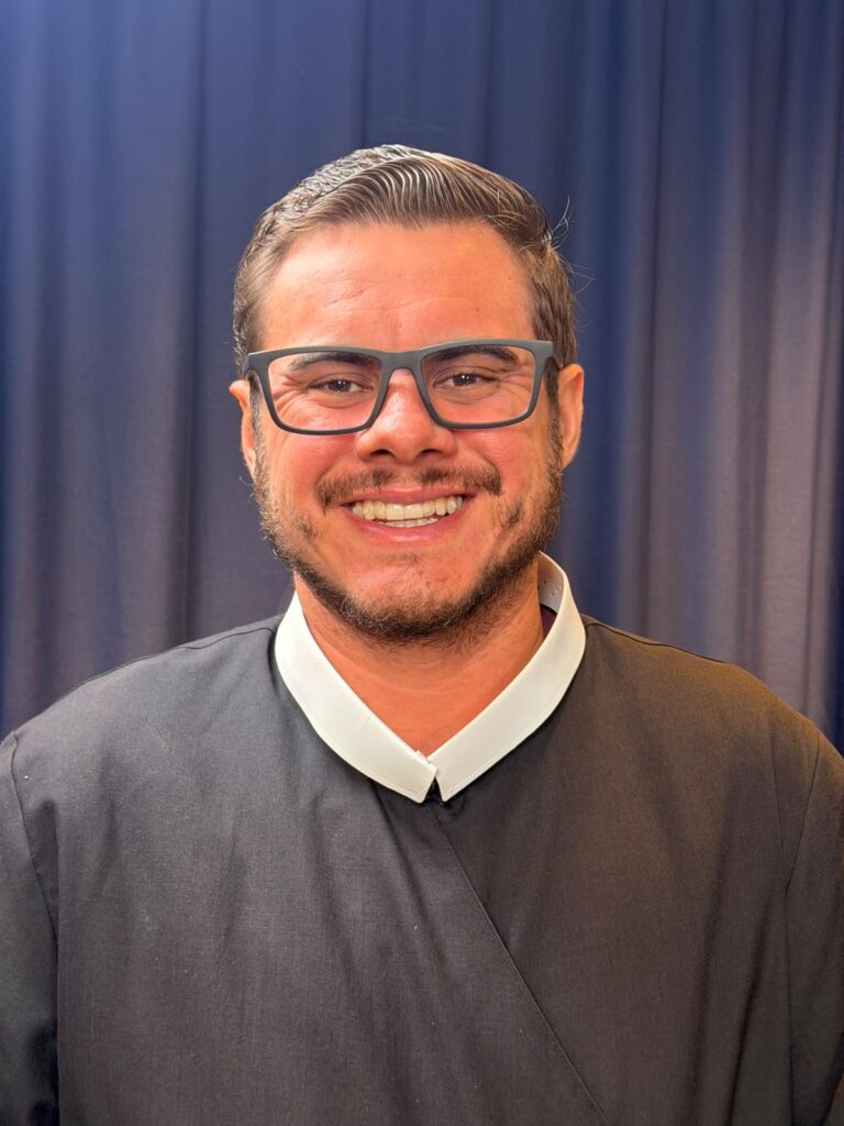 "Andrey Reimer de Melo, vestido com o hábito preto da Congregação Redentorista, está ajoelhado em oração diante de uma cruz de madeira rústica e uma pequena imagem de Nossa Senhora do Perpétuo Socorro. A cena evoca contemplação e serviço aos mais pobres, alinhado ao carisma de Santo Afonso de Ligório."