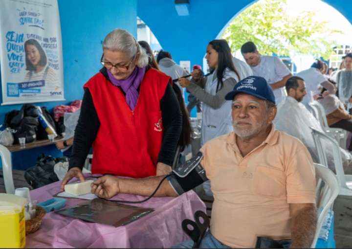 Voluntária da Pastoral da AIDS do Santuário do Rocio aferindo a pressão arterial de um cidadão. O momento ilustra o cuidado prático, a escuta e o acolhimento oferecidos durante a ação de saúde no Dia Lilás.